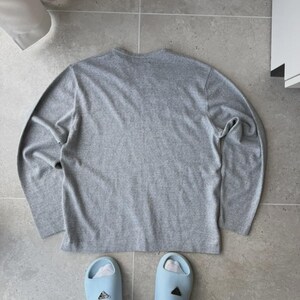 Op de afbeelding: Een grijs gem&ecirc;leerd shirt met lange mouwen en lichtblauwe slippers met witte sokken. Het shirt is effen en heeft een ronde hals. De slippers hebben een logo.