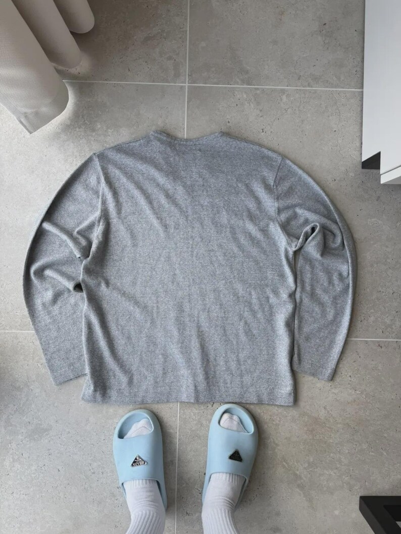 Op de afbeelding: Een grijs gem&ecirc;leerd shirt met lange mouwen en lichtblauwe slippers met witte sokken. Het shirt is effen en heeft een ronde hals. De slippers hebben een logo.
