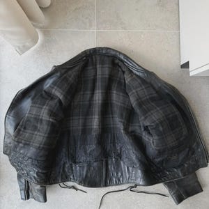 May include: Black leather jacket with a plaid lining. The jacket is open, revealing the interior lining. The jacket has a zipper closure and pockets. The leather appears worn, giving it a vintage look.