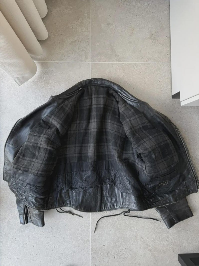 May include: Black leather jacket with a plaid lining. The jacket is open, revealing the interior lining. The jacket has a zipper closure and pockets. The leather appears worn, giving it a vintage look.