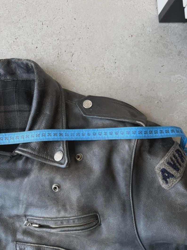 May include: Black leather jacket with a zipper closure and a small patch that reads "AVIA". The jacket is measured with a blue measuring tape. The jacket's shoulder width is approximately 18 inches.