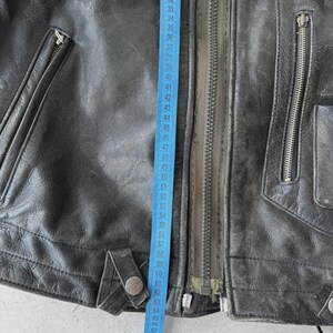 May include: Close-up of a black leather jacket with a silver zipper and multiple pockets. A blue measuring tape shows a length of approximately 27 to 73 inches. The jacket has a worn, vintage appearance.