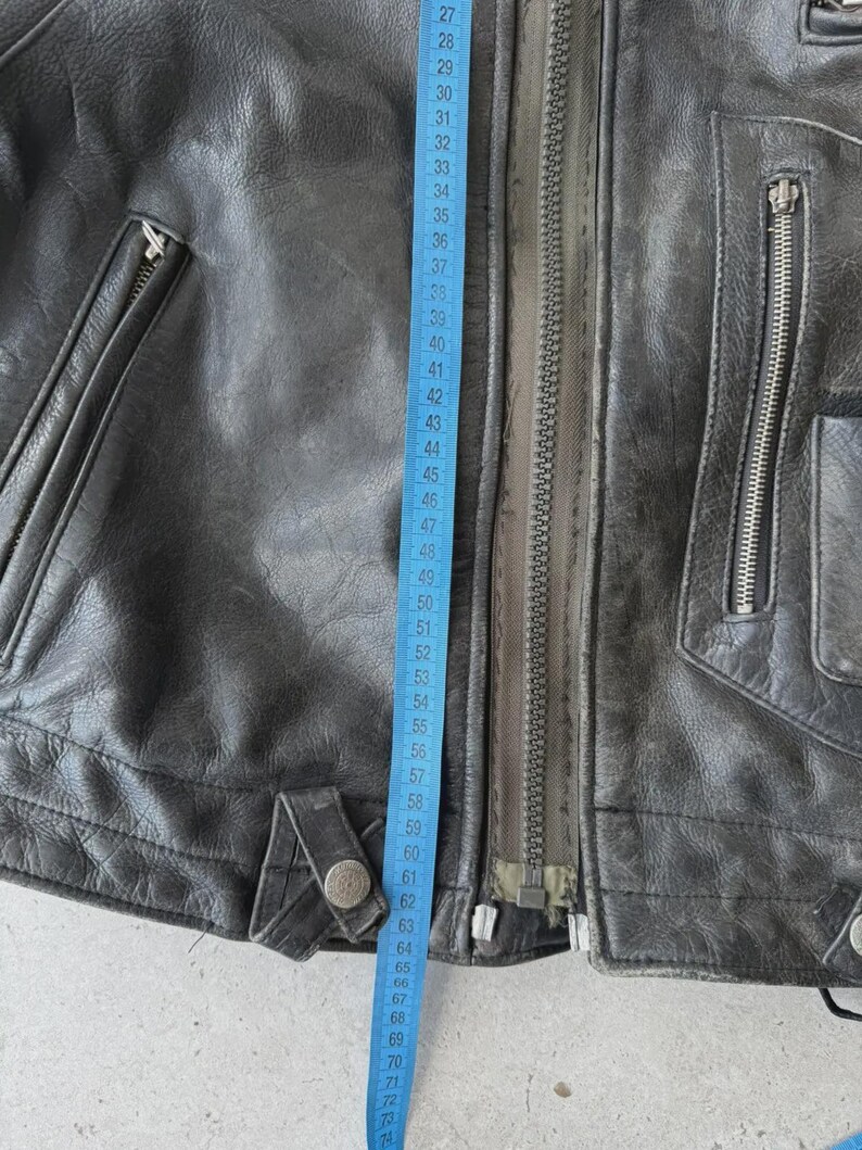 May include: Close-up of a black leather jacket with a silver zipper and multiple pockets. A blue measuring tape shows a length of approximately 27 to 73 inches. The jacket has a worn, vintage appearance.