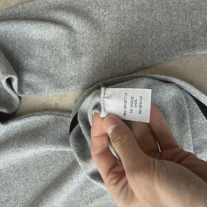 Op de afbeelding: Een close-up van een grijze trui met een zwart streepdetail. Het label van de trui toont de samenstelling: 50% merinowol, 50% acryl. De stof ziet er zacht en comfortabel uit.