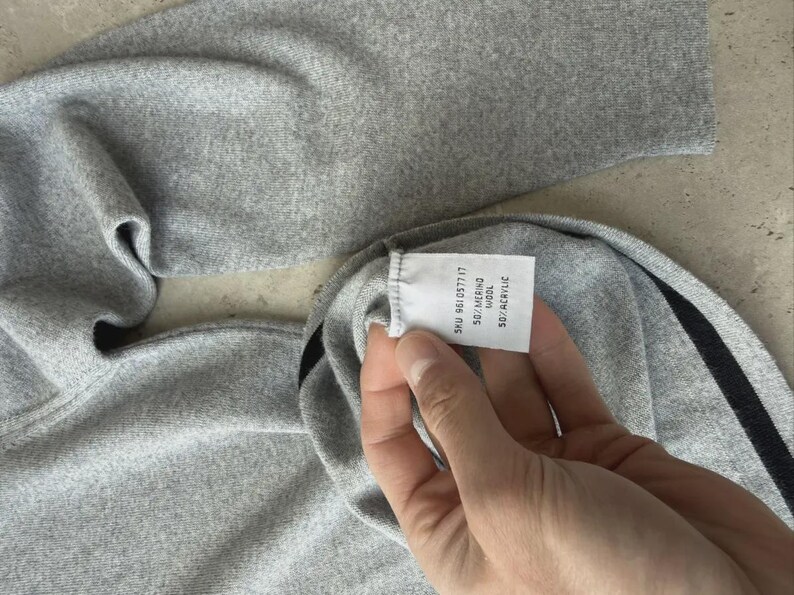 Op de afbeelding: Een close-up van een grijze trui met een zwart streepdetail. Het label van de trui toont de samenstelling: 50% merinowol, 50% acryl. De stof ziet er zacht en comfortabel uit.
