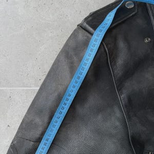 May include: Close-up of a black leather jacket with a blue measuring tape. The jacket has a classic design with a notched collar, shoulder epaulets, and a zipper closure. The measuring tape shows a length of approximately 28 inches.