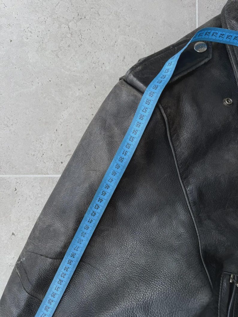 May include: Close-up of a black leather jacket with a blue measuring tape. The jacket has a classic design with a notched collar, shoulder epaulets, and a zipper closure. The measuring tape shows a length of approximately 28 inches.