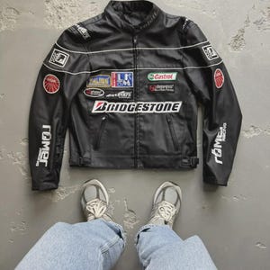 May include: Black racing jacket with multiple sponsor patches, including Bridgestone and Castrol. The jacket has white stripes and the word "Röme" on the sleeves. The jacket is displayed on a gray surface with a pair of gray sneakers and blue jeans.