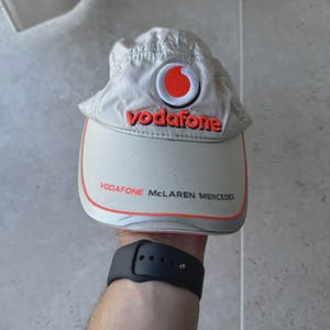 Könnte beinhalten: Beige Baseball Cap mit dem Vodafone-Logo in Rot und Weiß. Die Kappe hat einen gebogenen Schirm und den Text "VODAFONE McLaren MERCEDES" in Rot. Hergestellt aus einem leichten Material.