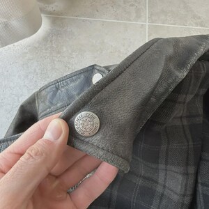 May include: Close-up of a dark gray leather jacket with a plaid lining. A silver button with the words "MOTORCYCLE CLUB AVIREX" is visible on the jacket's collar. The jacket appears worn and has a vintage aesthetic.