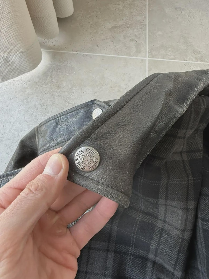 May include: Close-up of a dark gray leather jacket with a plaid lining. A silver button with the words "MOTORCYCLE CLUB AVIREX" is visible on the jacket's collar. The jacket appears worn and has a vintage aesthetic.