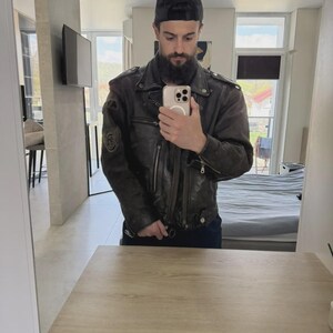 May include: A dark brown leather jacket with a black baseball cap. The jacket has a worn appearance with visible stitching and patches. The person is holding a phone in front of a mirror.