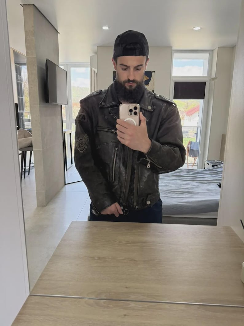 May include: A dark brown leather jacket with a black baseball cap. The jacket has a worn appearance with visible stitching and patches. The person is holding a phone in front of a mirror.