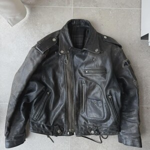 May include: A black leather motorcycle jacket with a zip-up front, multiple pockets, and a notched collar. The jacket has a worn appearance, with visible wear and tear. Features include shoulder epaulets and a small patch on the sleeve.