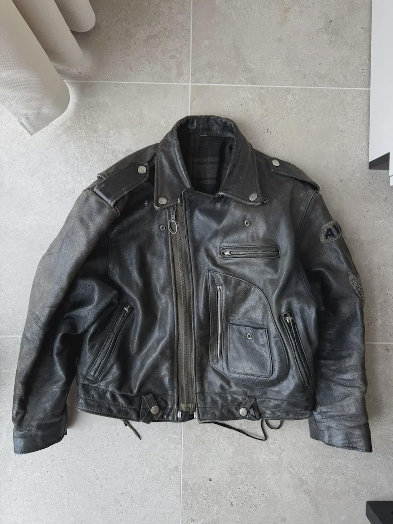 May include: A black leather motorcycle jacket with a zip-up front, multiple pockets, and a notched collar. The jacket has a worn appearance, with visible wear and tear. Features include shoulder epaulets and a small patch on the sleeve.