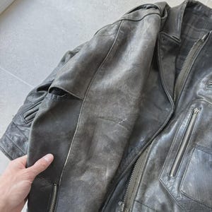 May include: A distressed gray leather jacket with a classic biker style. The jacket features a notched collar, multiple zippered pockets, and a front zipper closure. The leather shows signs of wear, adding to its vintage appeal.