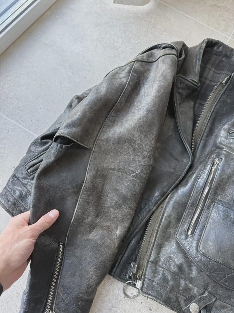 May include: A distressed gray leather jacket with a classic biker style. The jacket features a notched collar, multiple zippered pockets, and a front zipper closure. The leather shows signs of wear, adding to its vintage appeal.