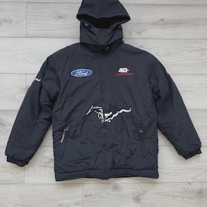 Op de afbeelding: Zwarte jas met capuchon met het Ford-logo en een wit Mustang-embleem. De jas heeft een ritssluiting, twee voorzakken en de tekst "40th Anniversary" in het rood. Ontworpen voor koud weer.