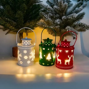 以下が含まれることがあります： 白、緑、赤の3つの装飾的なクリスマスランタン。白いランタンには雪の結晶の切り抜き、緑色のランタンには木の切り抜き、赤いランタンにはハートと鹿の切り抜きがあります。各ランタンにはハンドルと小さな蓋が付いています。