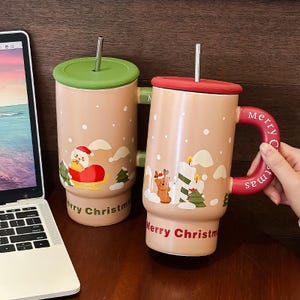 Può includere: Due tazze beige a tema natalizio con coperchi e cannucce. Una tazza presenta Babbo Natale su una slitta, l'altra una renna e delle candele. Entrambe le tazze hanno la scritta "Merry Christmas" e sono decorate con fiocchi di neve e alberi.