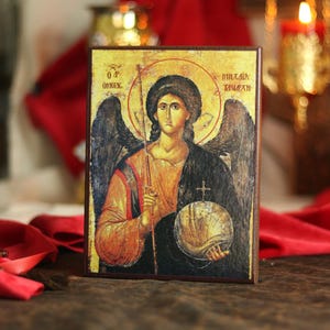 Puede incluir: Un icono rectangular de madera con una pintura religiosa de un ángel con alas oscuras, vestido con una túnica roja y negra. La figura sostiene un bastón fino y una esfera rematada con una cruz. Se observa texto griego en los lados.