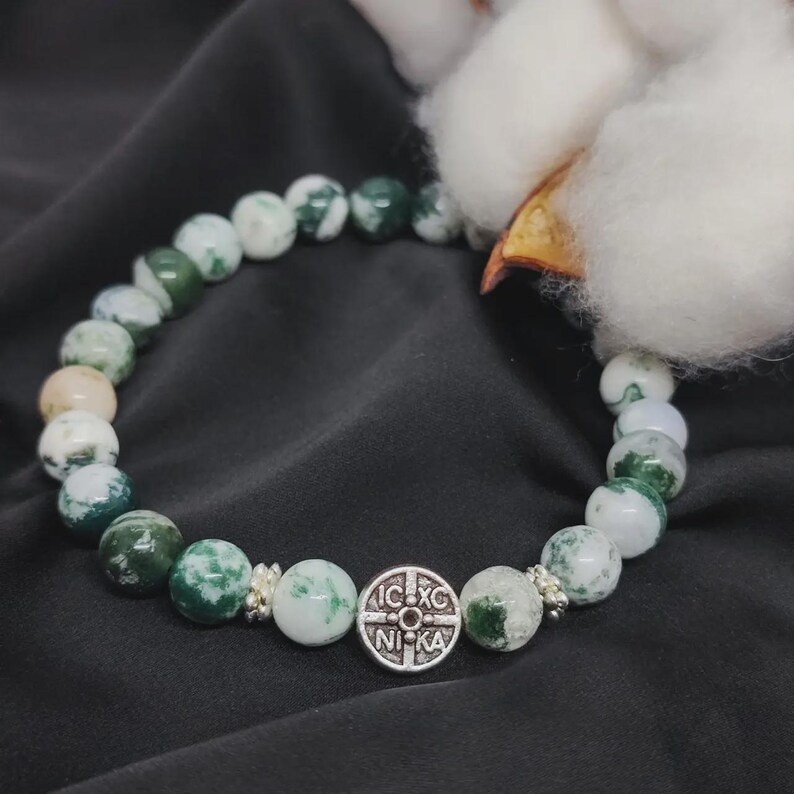May include: A beaded bracelet featuring round, green and white stone beads. A silver-toned charm with the text "IC XC NIKA" is centered on the bracelet. The bracelet is displayed on a dark gray surface.
