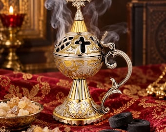 Può includere: Un incensiere in metallo con finitura oro e argento, caratterizzato da intricate incisioni a croce e sormontato da una croce decorativa. Il fumo si sprigiona dal coperchio traforato. L'oggetto presenta un manico curvo e una base ampia e stabile.