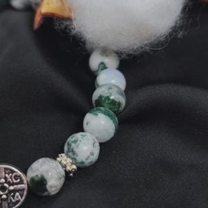 May include: A close-up of a bracelet featuring round, white and green moss agate beads. The bracelet has a silver-colored charm with an engraved design. The background is a dark, textured fabric.