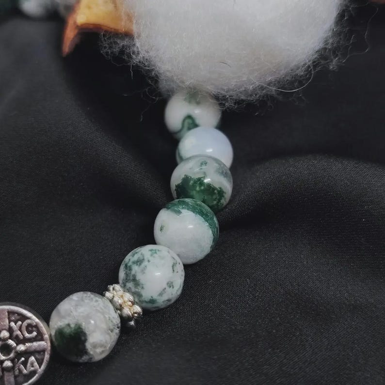 May include: A close-up of a bracelet featuring round, white and green moss agate beads. The bracelet has a silver-colored charm with an engraved design. The background is a dark, textured fabric.