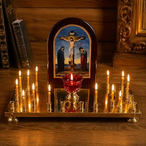 May include: A rectangular brass candle stand with multiple small lit candles and a central red glass votive holder. Behind the stand sits an arched wooden icon depicting the crucifixion of Jesus with two figures standing at the base of the cross.