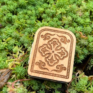 May include: A small, rectangular wooden box with a carved floral design on the lid. The box is light brown and sits on a bed of green plants. The floral design is a four-leaf clover shape with decorative swirls.