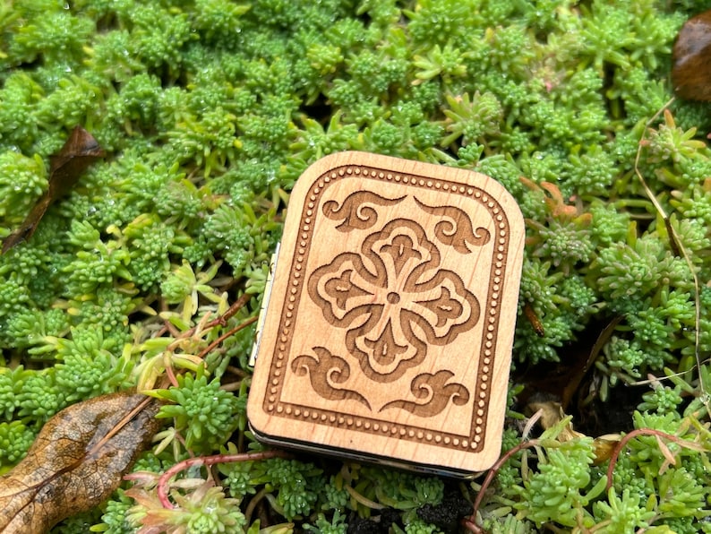 May include: A small, rectangular wooden box with a carved floral design on the lid. The box is light brown and sits on a bed of green plants. The floral design is a four-leaf clover shape with decorative swirls.