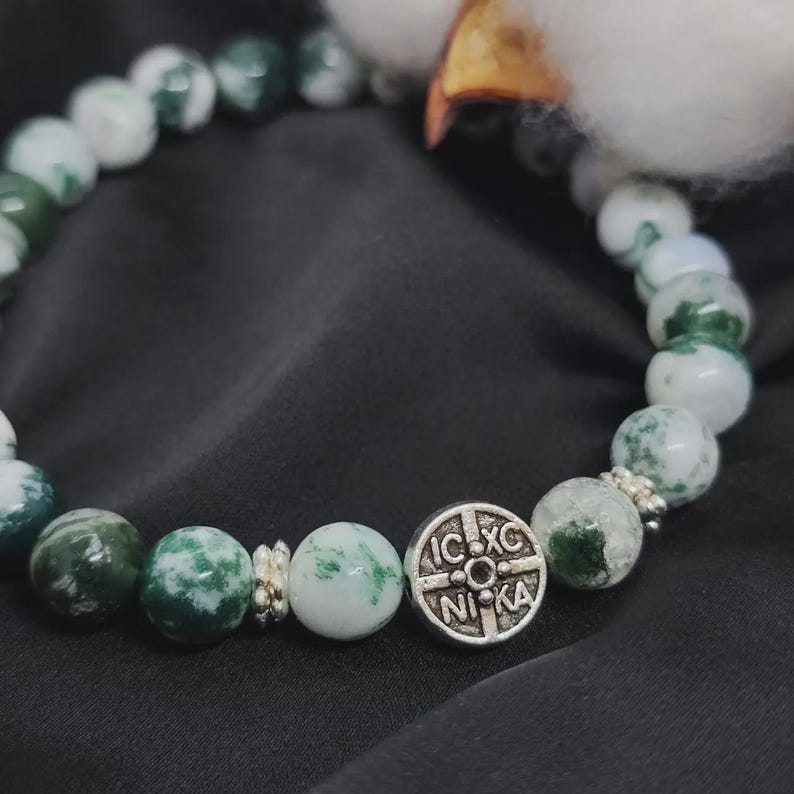 May include: A beaded bracelet featuring green and white speckled stones. A silver-colored charm with text is centered on the bracelet. The bracelet is displayed on a black surface.
