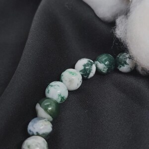 May include: A close-up of a bracelet featuring round beads. The beads are primarily white with green, moss-like patterns. The bracelet is set against a black background, with a fluffy white element in the upper right corner.