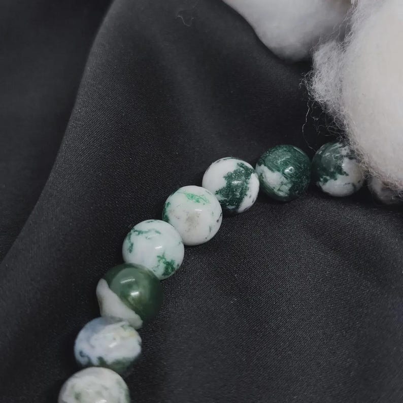 May include: A close-up of a bracelet featuring round beads. The beads are primarily white with green, moss-like patterns. The bracelet is set against a black background, with a fluffy white element in the upper right corner.
