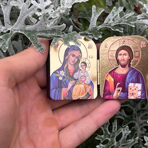 May include: Two small, rectangular religious icons with gold-colored frames. One depicts Mary and baby Jesus, the other Jesus. The icons are held in a hand against a background of green and white foliage.