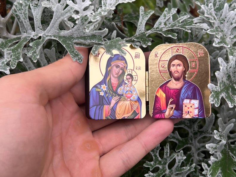 May include: Two small, rectangular religious icons with gold-colored frames. One depicts Mary and baby Jesus, the other Jesus. The icons are held in a hand against a background of green and white foliage.