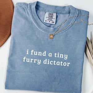 以下が含まれることがあります： 「i fund a tiny furry dictator」の白い文字が入った青色のComfort Colors Tシャツ。ペンダント付きのゴールドネックレスがシャツの上にかけられています。茶色の帽子と乾燥した植物が背景にあります。