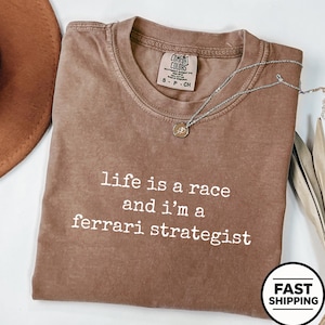 Op de afbeelding: Een bruin t-shirt met korte mouwen, een ronde hals en de witte opdruk "life is a race and i'm a ferrari strategist". Het shirt is voorzien van een Comfort Colors merklabel in de nek en een dunne zilverkleurige ketting.