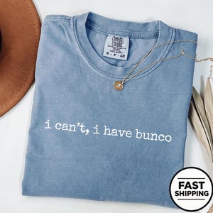 May include: A blue short-sleeve t-shirt with a crew neckline and the white text "i can't, i have bunco" printed on the front. The shirt features a Comfort Colors brand label inside the collar, indicating a size small, and is made of cotton.
