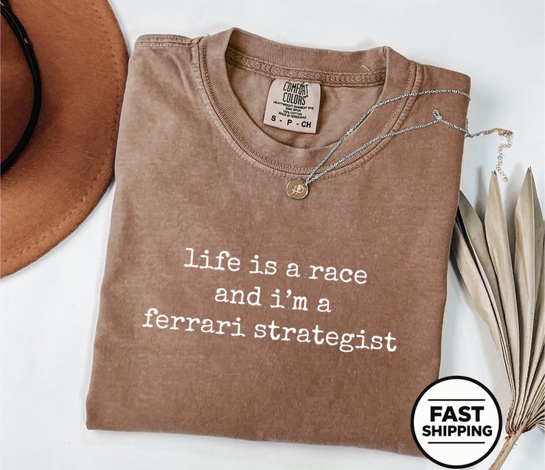 Op de afbeelding: Een bruin t-shirt met korte mouwen, een ronde hals en de witte opdruk "life is a race and i'm a ferrari strategist". Het shirt is voorzien van een Comfort Colors merklabel in de nek en een dunne zilverkleurige ketting.