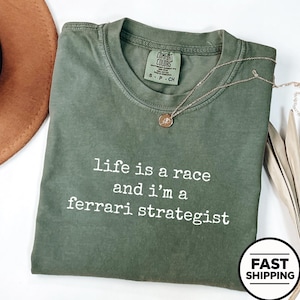 Op de afbeelding: Een olijfgroen t-shirt met korte mouwen, een ronde hals en de witte opdruk "life is a race and i'm a ferrari strategist". Het shirt is voorzien van een Comfort Colors-merklabel in de binnenkant van de hals en is gemaakt van katoen.