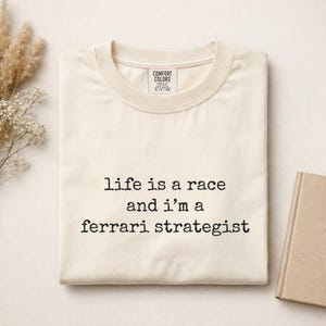 Op de afbeelding: Een cr&egrave;mekleurig t-shirt met korte mouwen en een ronde hals. Het t-shirt heeft zwarte tekst op de voorkant gedrukt die luidt: life is a race and i'm a ferrari strategist. Het label in de nek aan de binnenzijde toont de merknaam Comfort Colors.