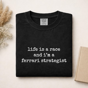 Op de afbeelding: Een zwart t-shirt met ronde hals en de witte tekst "life is a race and i'm a ferrari strategist" op de voorzijde. Het t-shirt heeft een Comfort Colors merklabel dat zichtbaar is aan de binnenkant van de halslijn.