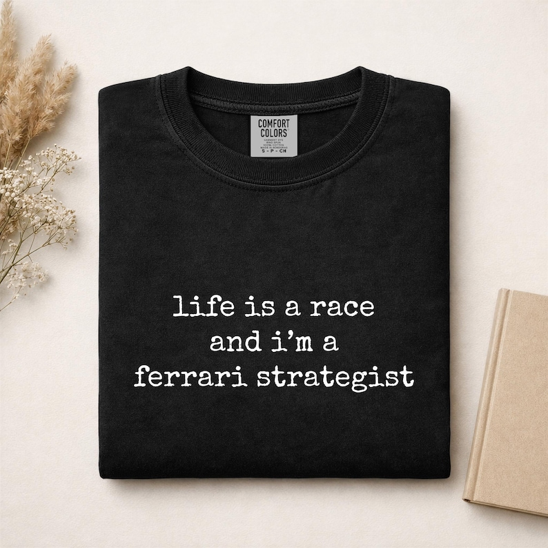 Op de afbeelding: Een zwart t-shirt met ronde hals en de witte tekst "life is a race and i'm a ferrari strategist" op de voorzijde. Het t-shirt heeft een Comfort Colors merklabel dat zichtbaar is aan de binnenkant van de halslijn.