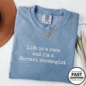 Op de afbeelding: Een blauw t-shirt met korte mouwen en de witte tekst "life is a race and i'm a ferrari strategist" op de voorkant. Het shirt heeft een ronde hals en een Comfort Colors merklabel in de kraag, gecombineerd met een gouden ketting.
