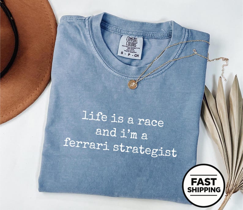 Op de afbeelding: Een blauw t-shirt met korte mouwen en de witte tekst "life is a race and i'm a ferrari strategist" op de voorkant. Het shirt heeft een ronde hals en een Comfort Colors merklabel in de kraag, gecombineerd met een gouden ketting.