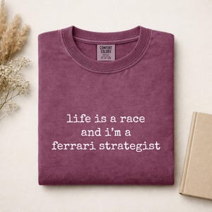 Op de afbeelding: Een donkerbordeauxrood t-shirt met ronde hals, een geribbelde kraag en een Comfort Colors merklabel. Op de voorkant staat witte tekst: life is a race and i'm a ferrari strategist. Het shirt heeft een zachte, vervaagde textuur en korte mouwen.