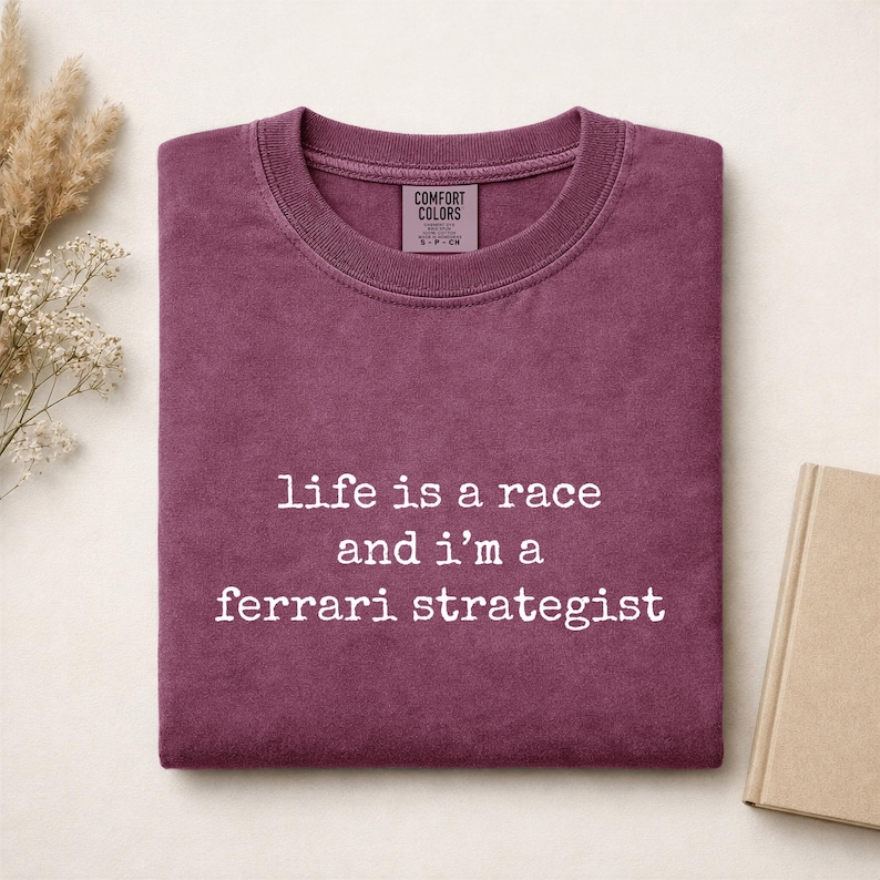 Op de afbeelding: Een donkerbordeauxrood t-shirt met ronde hals, een geribbelde kraag en een Comfort Colors merklabel. Op de voorkant staat witte tekst: life is a race and i'm a ferrari strategist. Het shirt heeft een zachte, vervaagde textuur en korte mouwen.