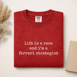 Op de afbeelding: Een rood t-shirt met ronde hals en de witte tekst "life is a race and i'm a ferrari strategist" op de voorkant. Het shirt heeft een Comfort Colors merklabel aan de binnenkant van de kraag en is gemaakt van een effen rode stof.